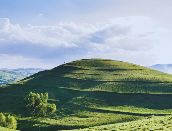 colina cubierta de verde hierba con árboles y bajo un cielo azul con nubes