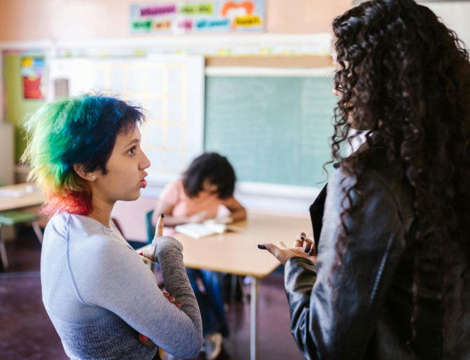 Dos alumnas mantienen una discusión mientras observan a otra en su pupitre.