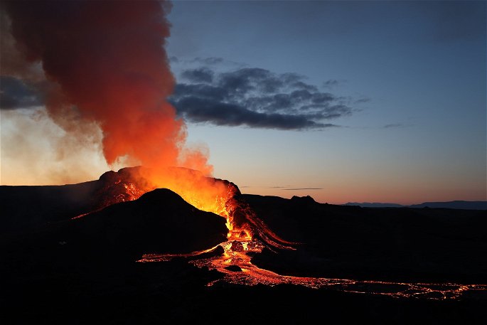 Un volcán en erupción durante el anochecer, desprendiendo gases y un río de lava