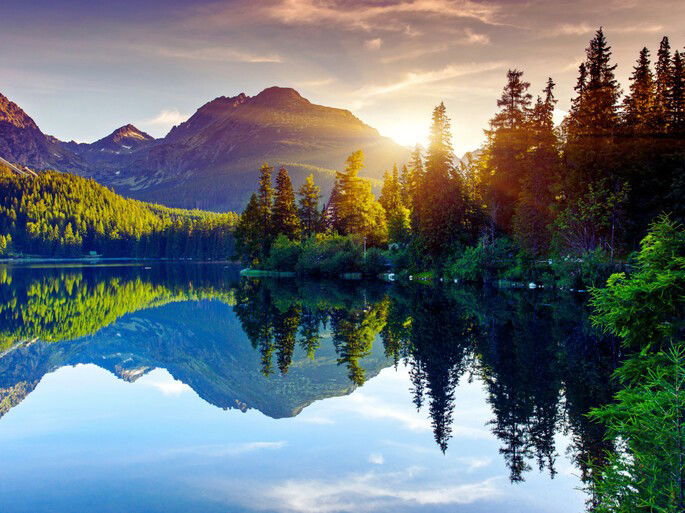 Un bosque montañoso rodeando un lago que refleja el mismo y el cielo nublado, en un atardecer.