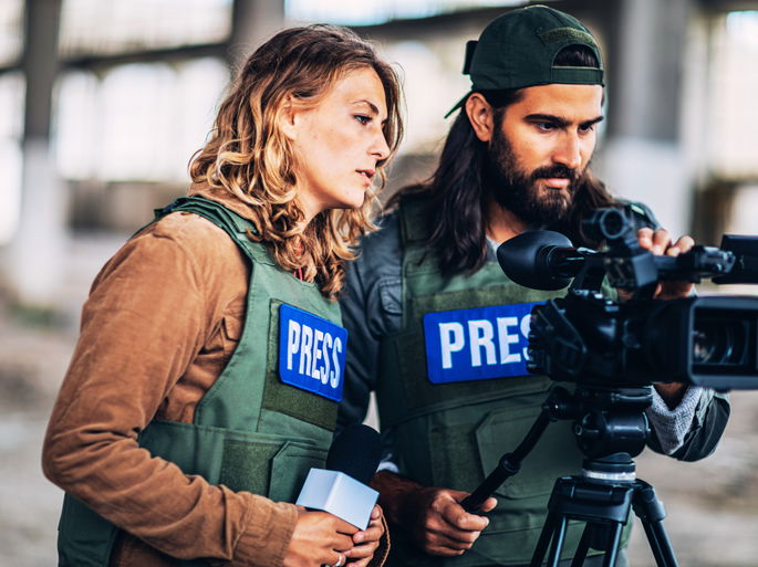 Dos periodistas graban con un cámara, mientras llevan chalecos de prensa para ser reconocidos.