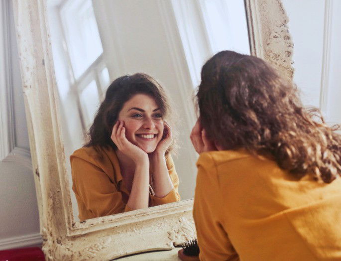 chica con camisa amarillo mostaza observa su reflejo en un espejo con marco blanco y antiguo